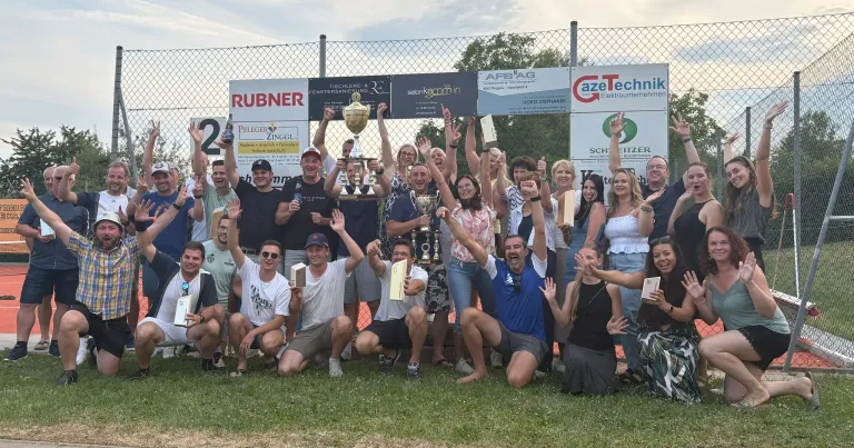 Eine Gruppe von Menschen feiert mit Trophäen auf einem Feld, einige sitzend und andere stehend. Hinter ihnen steht ein Zaun mit Bannern und Werbetafeln.