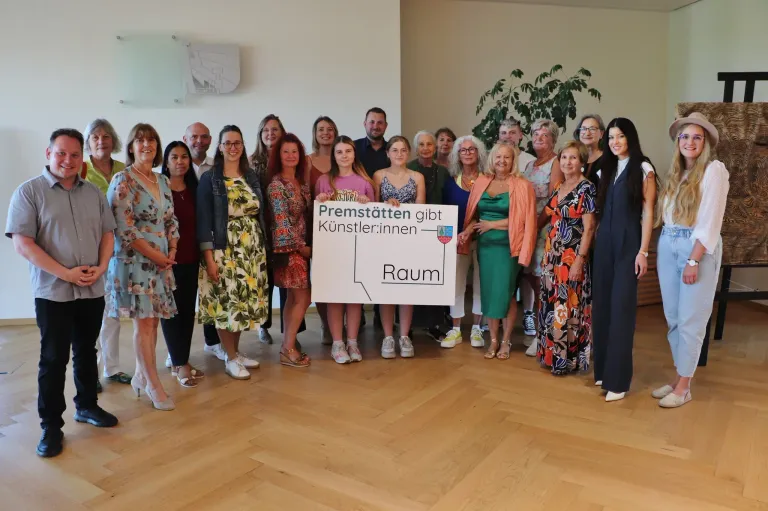 Eine Gruppe von Menschen steht zusammen und hält ein Schild mit der Aufschrift 'Premstatten gibt Künstlern Raum'. Sie sind alle in verschiedenen Outfits gekleidet und einige tragen Brillen. Sie lächeln alle und posieren für ein Foto.