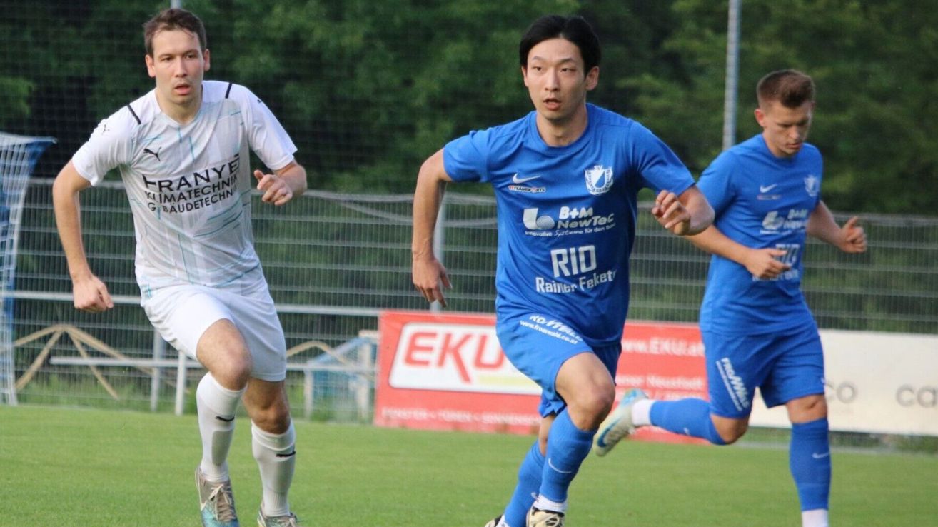 Ein Fußballspieler in einem blauen Trikot sprintet in Richtung Tor, während zwei weitere folgen. Das Spielfeld ist eingezäunt und ein rotes Banner mit EKU ist im Hintergrund sichtbar.