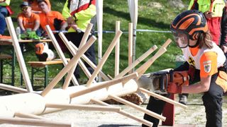 Ein Kettensägenwettbewerb ist im Gange, bei dem ein Teilnehmer eine Kettensäge benutzt, um Holzstämme zu schneiden. Zuschauer und Offizielle sind anwesend und beobachten die Veranstaltung.