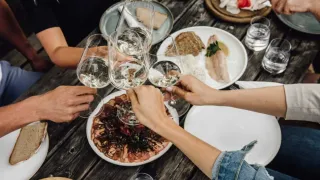 Eine Gruppe von Menschen sitzt um einen Holztisch herum und hält Weingläser. Es gibt verschiedene Teller mit Essen, einschließlich Fleisch, Fisch und Gemüse. Eine Flasche Wein steht auf dem Tisch.
