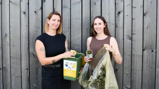 Zwei Frauen stehen lächelnd vor einer Holzwand. Eine hält einen grünen Recyclingbehälter, die andere einen Plastikbeutel. Sie scheinen für ein Foto zu posieren.