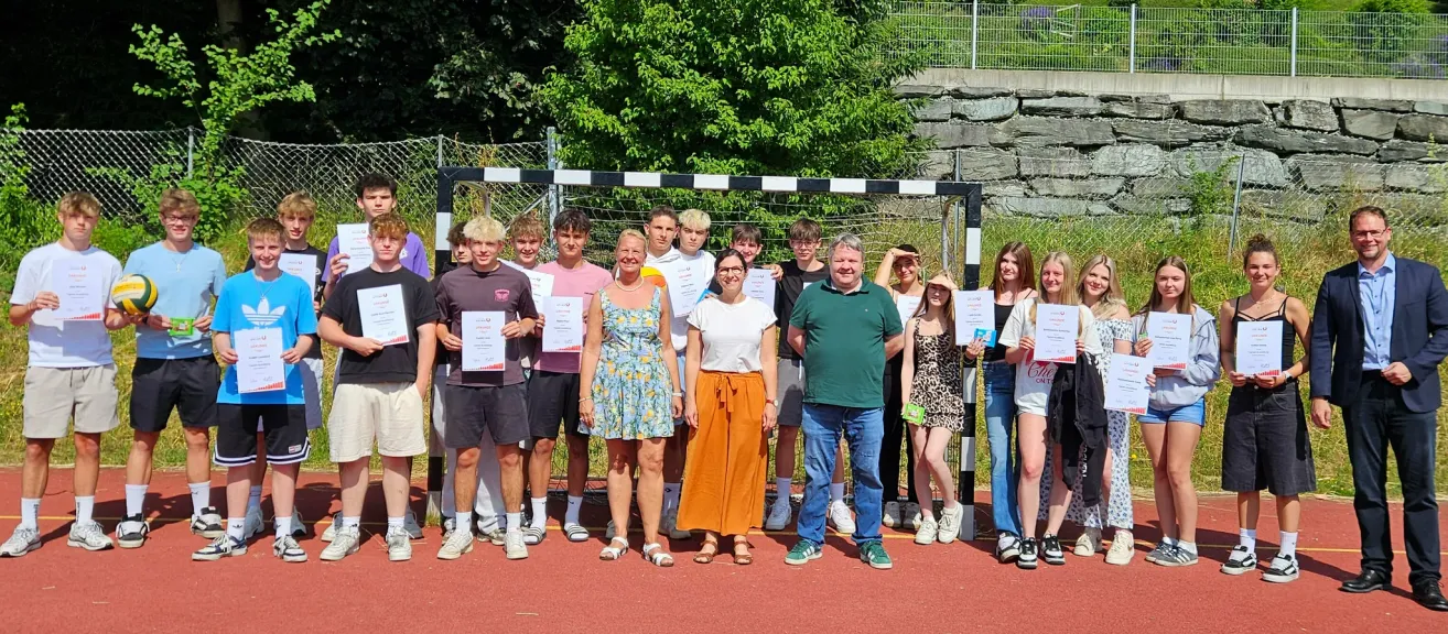 Eine Gruppe von Menschen, darunter junge Männer und Frauen, steht vor einem Fußballtor. Sie halten Urkunden in den Händen. Dahinter befindet sich ein Zaun mit einigen Pflanzen und Bäumen.