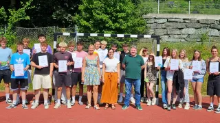 Eine Gruppe von Menschen, darunter junge Männer und Frauen, steht vor einem Fußballtor. Sie halten Urkunden in den Händen. Dahinter befindet sich ein Zaun mit einigen Pflanzen und Bäumen.