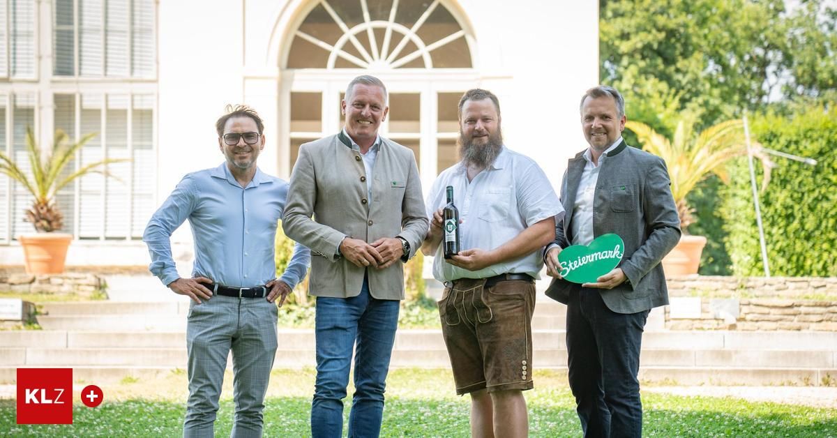 Vier Männer stehen vor einem Gebäude. Die mittleren beiden halten eine Flasche und ein grünes herzförmiges Schild. Der Hintergrund zeigt ein weißes Bauwerk mit einem bogenförmigen Fenster.