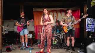 Eine Band tritt auf der Bühne auf, eine Frau singt. Zwei Männer spielen Gitarre, ein anderer Schlagzeug. Ein Banner steht dahinter, und verschiedene Geräte sind auf der Bühne.