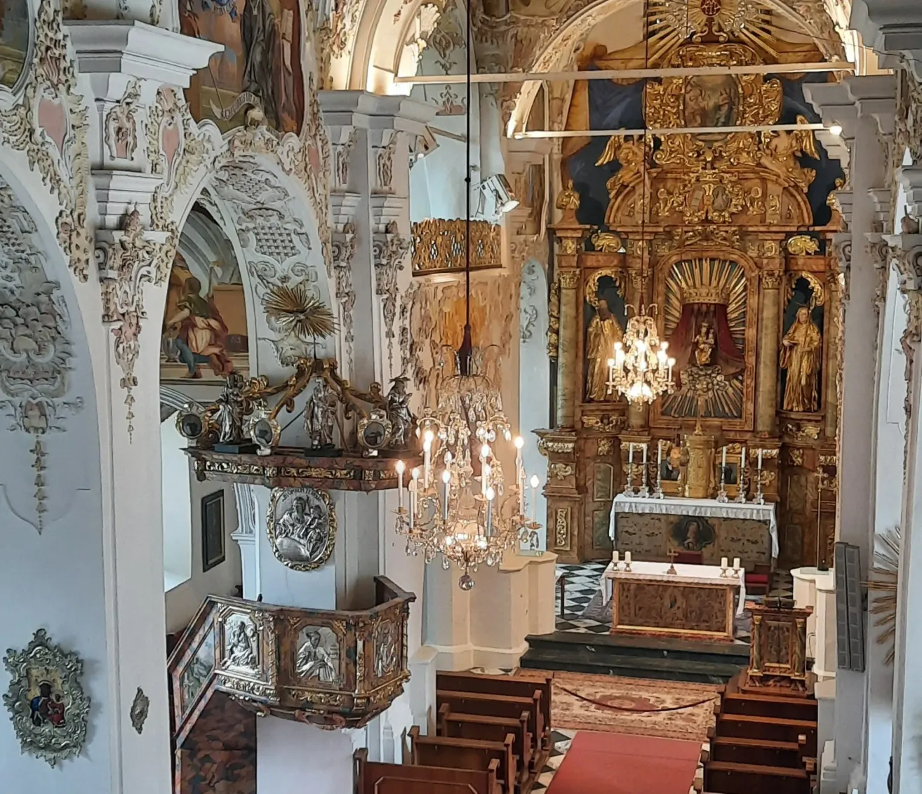 Ein reich verzierter Kircheninnenraum mit aufwendigen goldenen Altaraufsätzen, kunstvollen Schnitzereien und hängenden Kronleuchtern. Der Altar hat Statuen und Kerzen, und die Kirchenbänke sind auf beiden Seiten angeordnet.