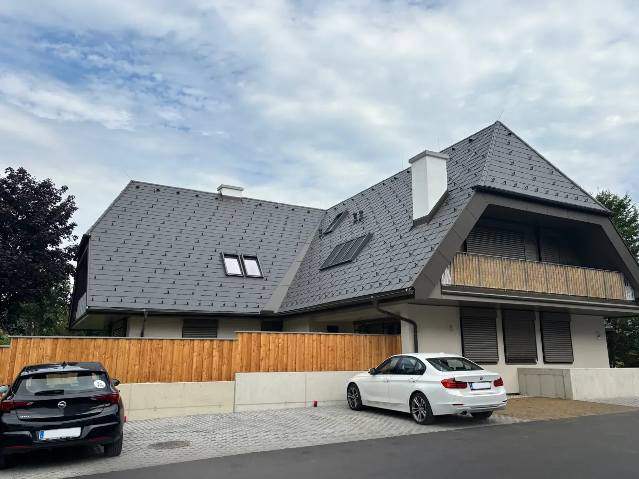 Ein modernes Haus mit grauem Dach und Balkon hat zwei Autos vor der Tür geparkt, eines weiß und eines schwarz, unter einem teilweise bewölkten Himmel.