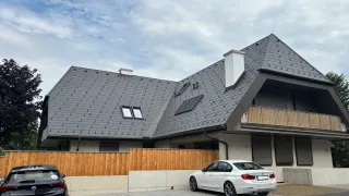 Ein modernes Haus mit grauem Dach und Balkon hat zwei Autos vor der Tür geparkt, eines weiß und eines schwarz, unter einem teilweise bewölkten Himmel.