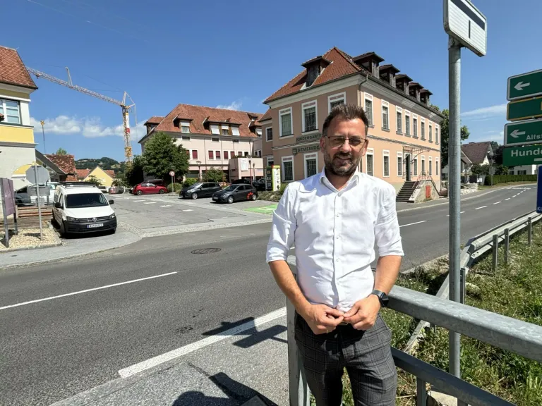 Ein Mann in einem weißen Hemd und einer Brille steht lächelnd auf einer Straße. Hinter ihm sind Gebäude, geparkte Autos und ein Kran zu sehen.