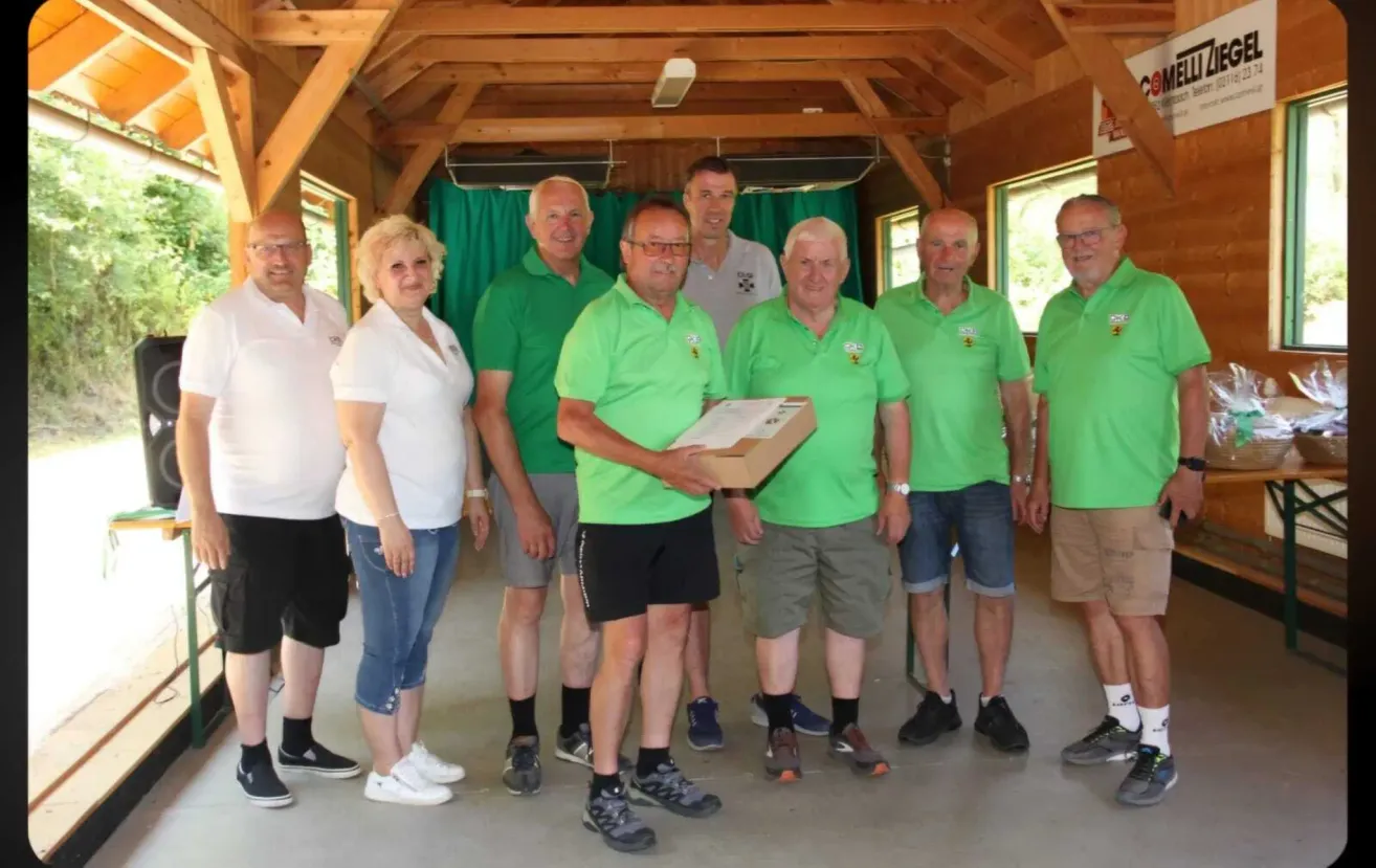 Eine Gruppe von Personen in grünen Polo-Shirts steht in einem hölzernen Gebäude. Ein Mann hält eine Box, während andere lächeln. Die Umgebung ist drinnen mit Holzbalken an der Decke.