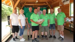 Eine Gruppe von Personen in grünen Polo-Shirts steht in einem hölzernen Gebäude. Ein Mann hält eine Box, während andere lächeln. Die Umgebung ist drinnen mit Holzbalken an der Decke.