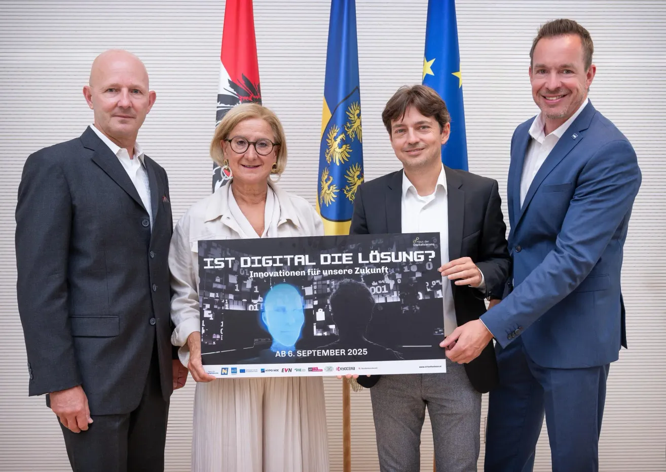 Vier Personen stehen lächelnd und halten ein Schild. Der Text auf dem Schild lautet 'IST DIGITAL DIE LÖSUNG?' mit dem Datum 6. September 2025. Dahinter sind drei Flaggen zu sehen.