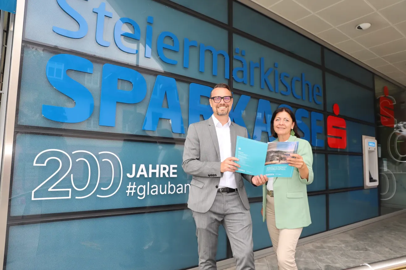 Zwei Personen stehen vor einem Gebäude mit dem Wort 'Sparkasse' darauf. Sie lächeln und halten eine Mappe. Der Mann trägt eine Brille und einen Anzug, und die Frau trägt einen grünen Blazer und Ohrringe. Das Gebäude hat Glaswände.