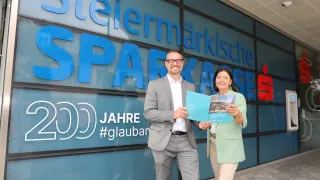 Zwei Personen stehen vor einem Gebäude mit dem Wort 'Sparkasse' darauf. Sie lächeln und halten eine Mappe. Der Mann trägt eine Brille und einen Anzug, und die Frau trägt einen grünen Blazer und Ohrringe. Das Gebäude hat Glaswände.