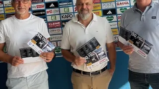 Drei Männer stehen vor einer Wand mit verschiedenen Werbeplakaten und halten einen Flyer mit der Aufschrift '50'. Sie alle lächeln und scheinen für ein Foto zu posieren.