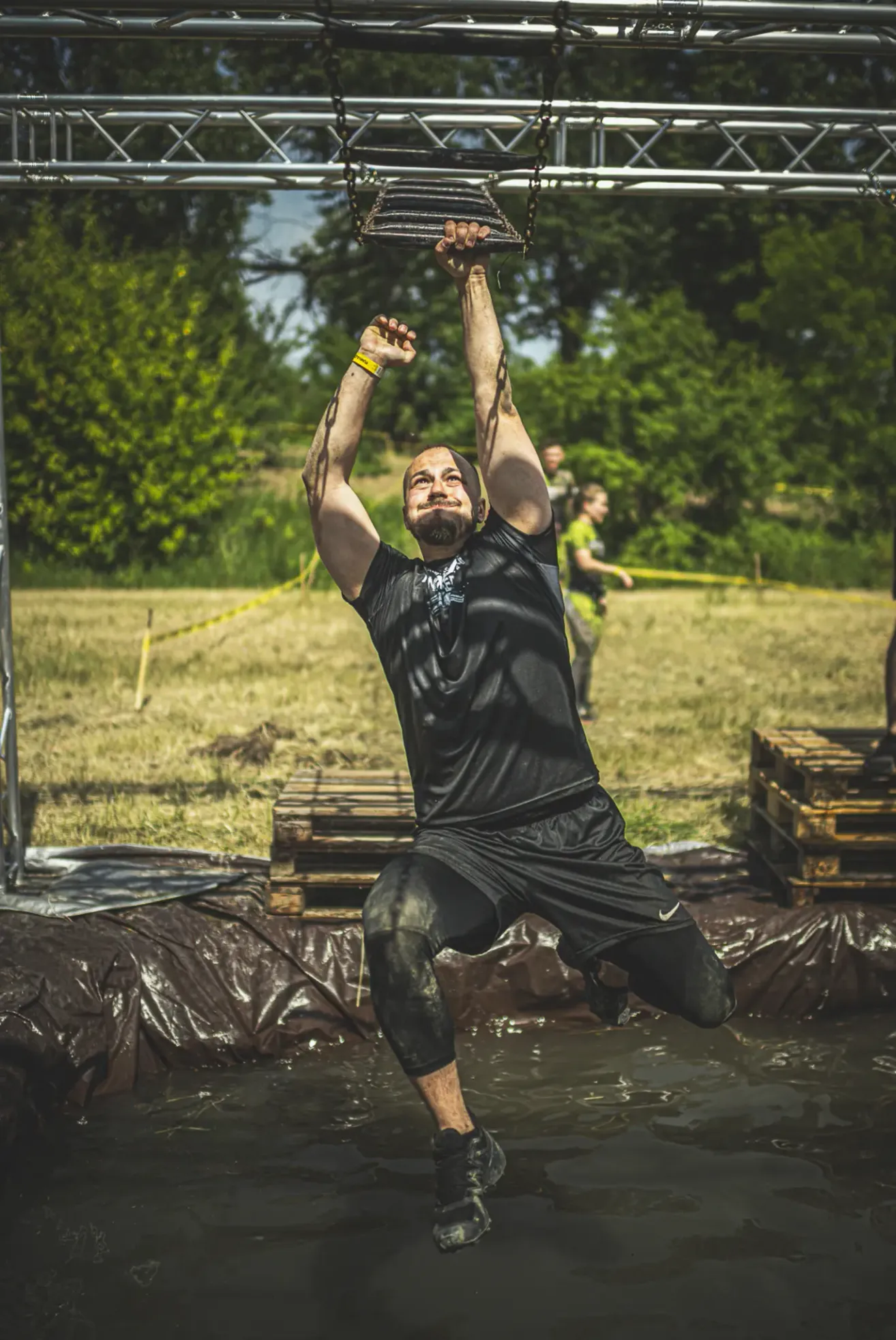 Ein Mann führt ein Klimmzug-Training an einer Stange in einem schlammigen Bereich durch. Er trägt ein schwarzes Outfit mit Nike-Logo. Hinter ihm stehen Holzpaletten. In der Ferne gibt es Bäume und zwei Personen, die gehen.