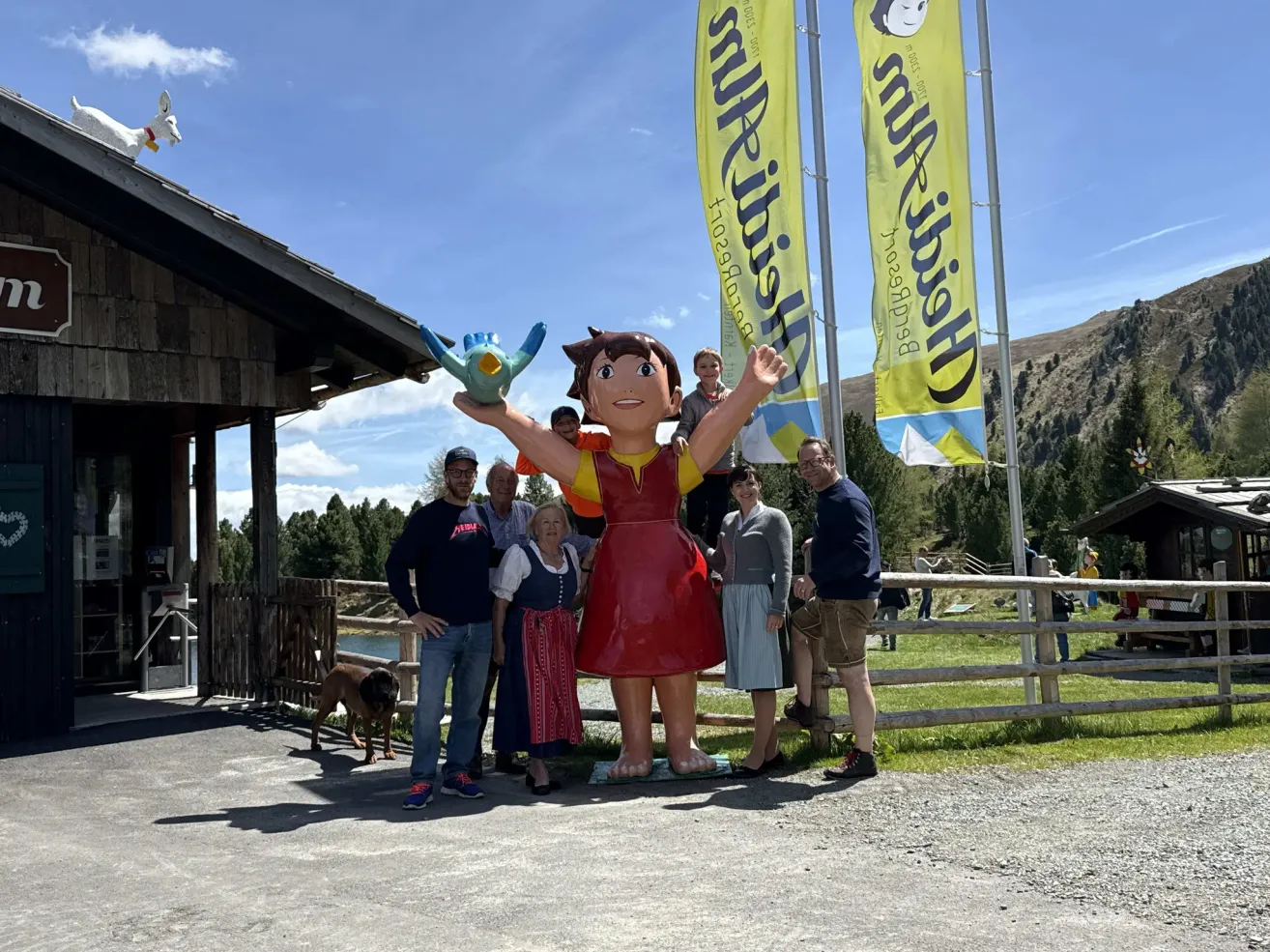Eine Gruppe von Menschen steht in der Nähe einer großen Heidi-Statue, mit einem Hund, einem Gebäude und einem Zaun im Hintergrund. Im Hintergrund befinden sich Banner mit dem Wort Heidi.