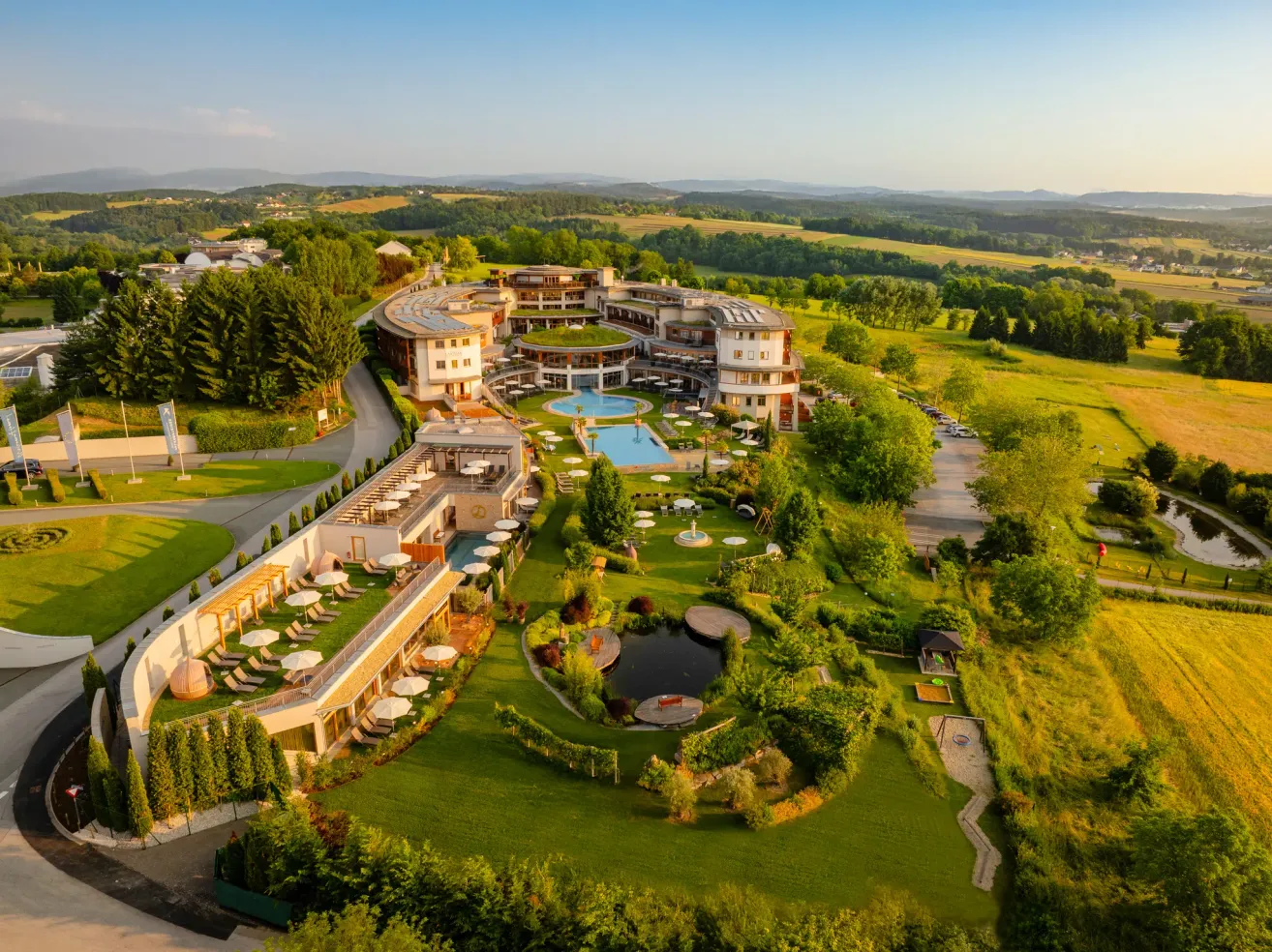 Luftaufnahme eines Resorts mit einem großen Schwimmbad, umgeben von grünem Rasen und Bäumen, mit Bergen in der Ferne.