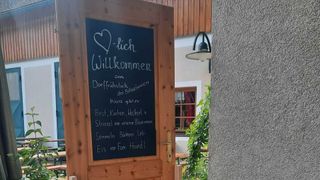 Eine Holztür mit einem Kreidetafelschild, das Besucher mit Frühstücksoptionen, Brot, Kuchen, Weckherl und Striezel von Bäckern Lieb sowie Eis von Fam. Heindl willkommen heißt.