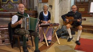 Drei Personen sitzen in einem Raum, wahrscheinlich einer Kirche, in traditioneller Kleidung. Ein Mann spielt Akkordeon, ein anderer Gitarre, und eine Frau steht in der Nähe.