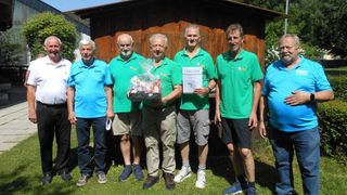 Eine Gruppe von Männern steht draußen, drei in grünen Shirts und einer hält ein Zertifikat. Sie lächeln und scheinen für ein Foto zu posieren.