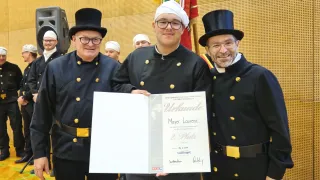 Ein Mann, der einen Zertifikat hält, steht zwischen zwei anderen in schwarzen Uniformen mit Zylinderhüten. Das Zertifikat trägt die Aufschrift 'Urkunde' und der Name des Mannes ist Mayer Laurenz. Sie stehen vor einer gemusterten Wand.