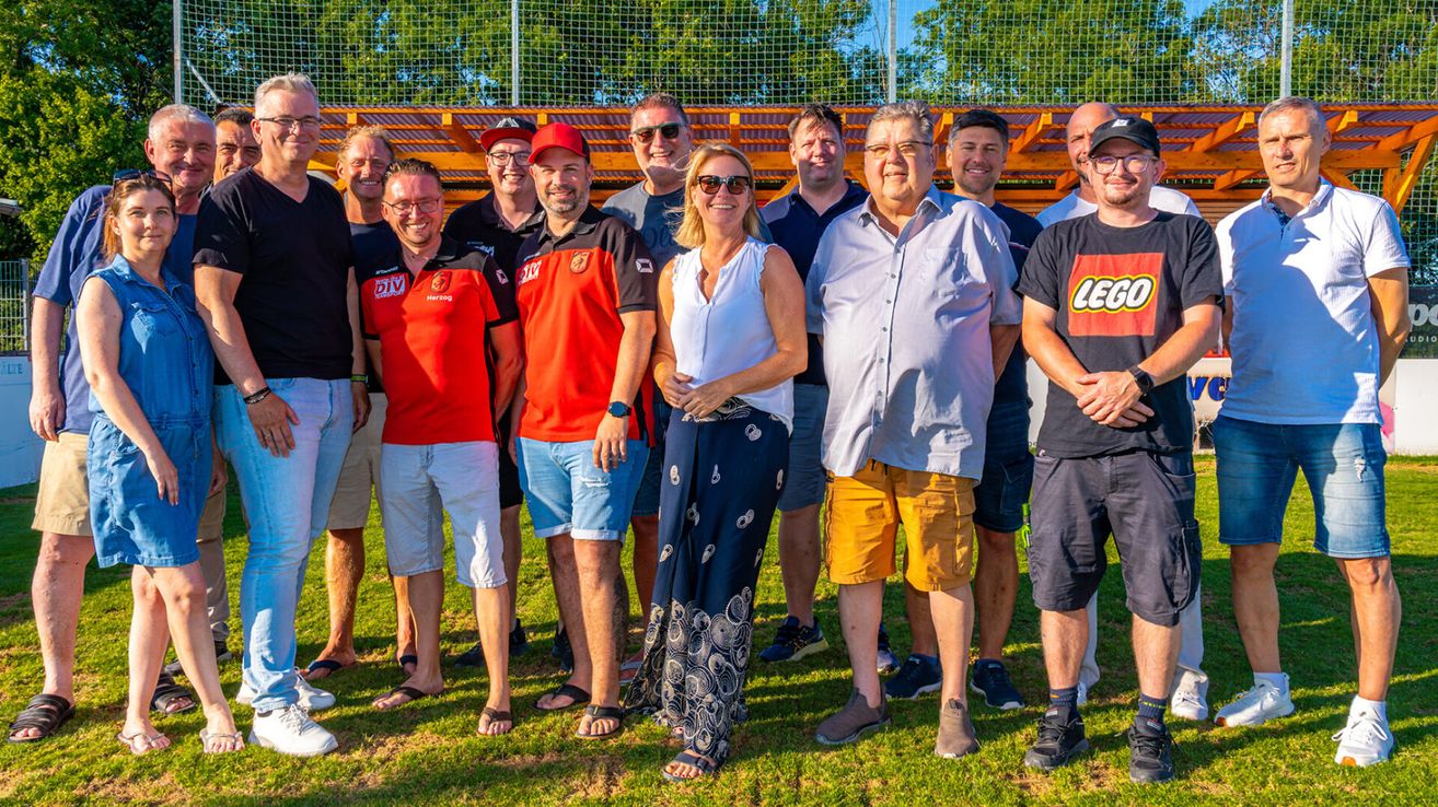Eine Gruppe von Menschen steht zusammen, lächelt und posiert für ein Foto. Sie befinden sich auf einem Rasenfeld mit einem Netzzaun im Hintergrund. Einige tragen Brillen und Sonnenbrillen.