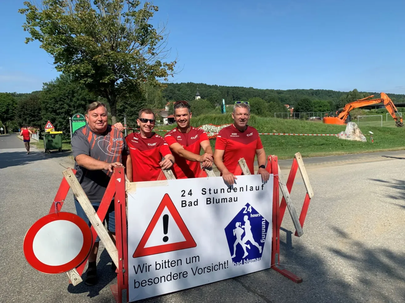 Vier Männer stehen hinter einem Schild, auf dem 24-Stunden-Marathon Bad Blumau steht. Sie tragen rote Hemden. Hinter ihnen befindet sich eine Baustelle mit Baumaschinen.