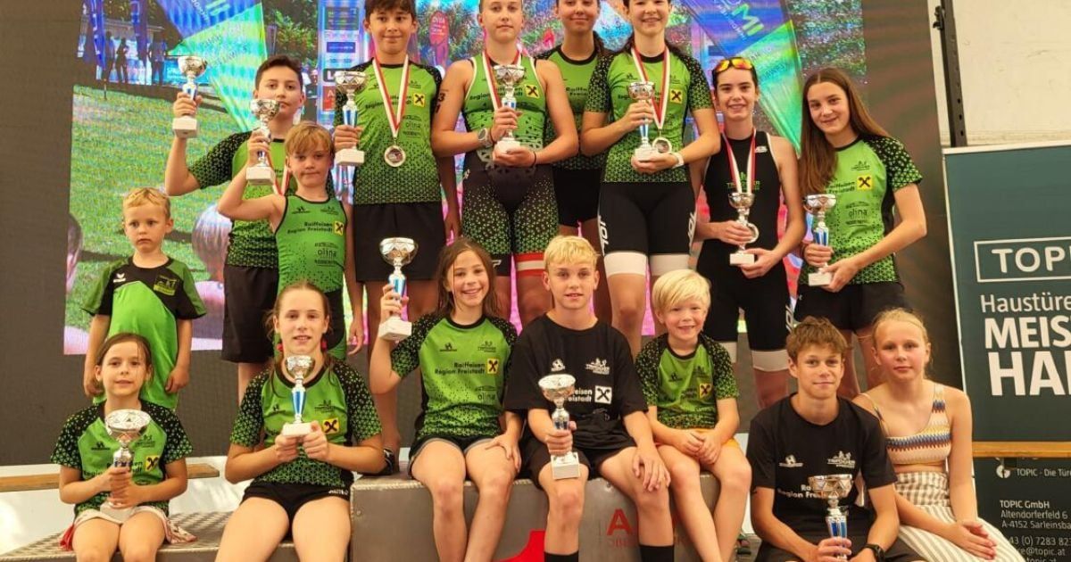 Eine Gruppe junger Athleten, gekleidet in grünen und schwarzen Uniformen, halten Trophäen und Medaillen auf einer Bühne, mit einem bunten Hintergrund hinter ihnen.
