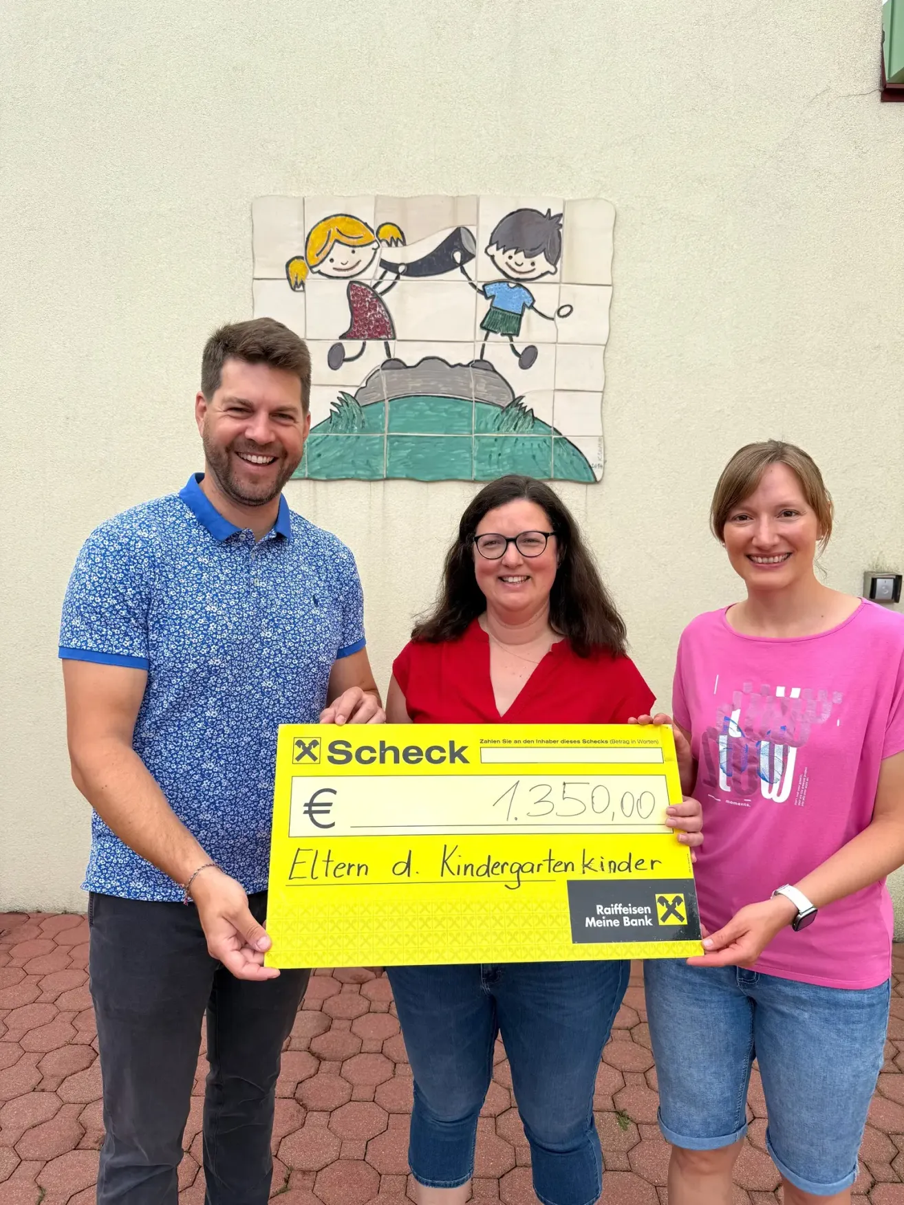 Drei Personen stehen vor einer Wandmalerei und halten einen gelben Scheck mit der Aufschrift 'Scheck' für 1.350,00 Euro, vermutlich für Eltern von Kindergartenkindern.