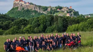 Eine Gruppe von Menschen, die in Schwarz und Rot gekleidet sind, posiert auf einer Wiese mit zwei Motorrädern vor ihnen, mit einer Burg auf einem Felsen im Hintergrund.