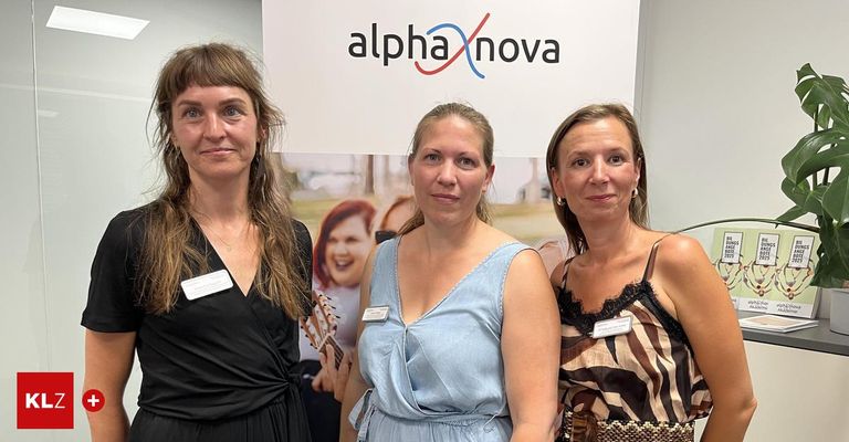 Drei Frauen posieren für ein Foto vor einem Alpha Nova-Schild. Eine hält eine Gitarre, alle tragen Namensschilder.