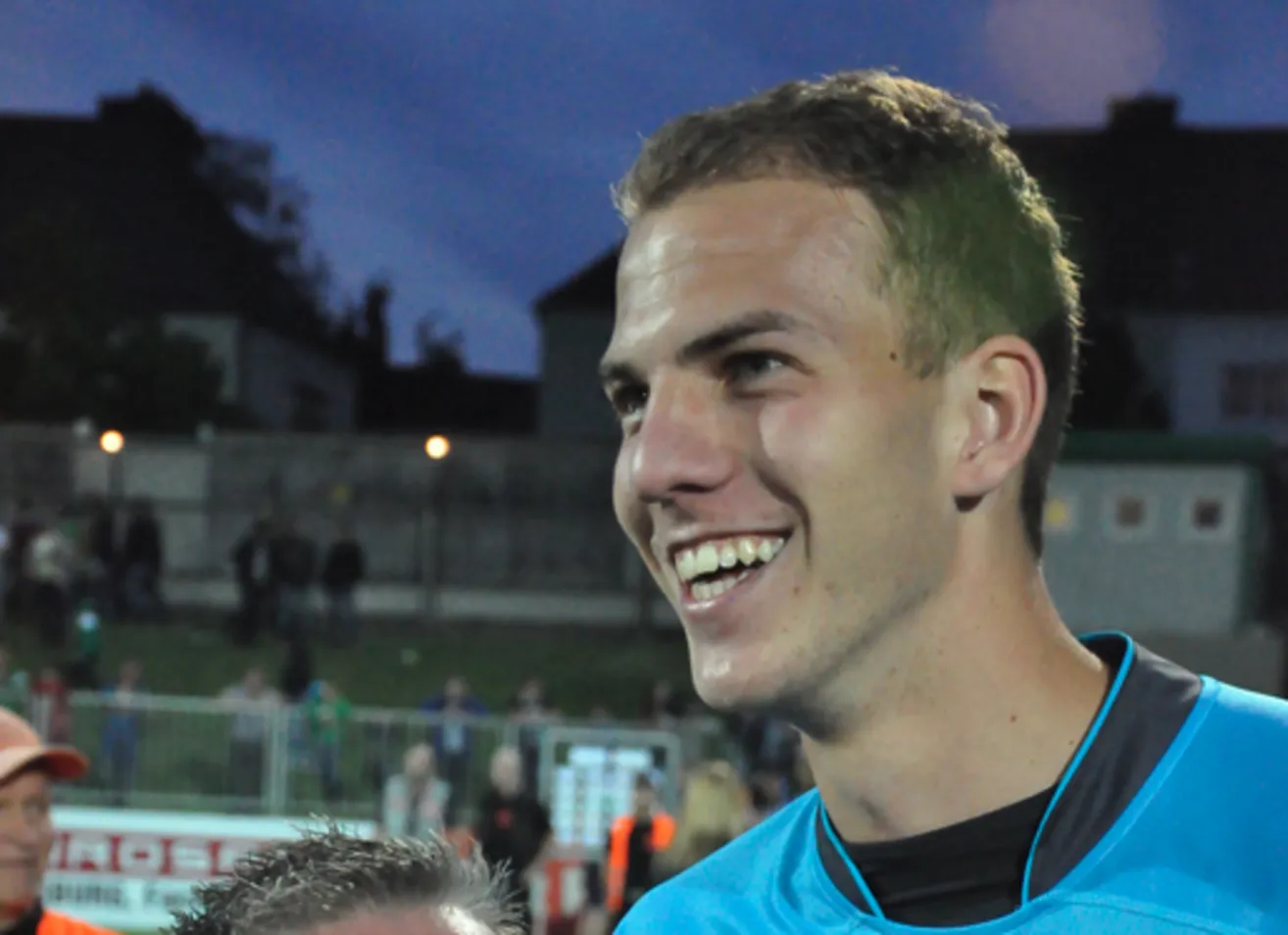 Ein lächelnder männlicher Fußballspieler steht nachts auf dem Feld und trägt ein blaues Trikot. Im Hintergrund steht eine Menschenmenge hinter einem Zaun.