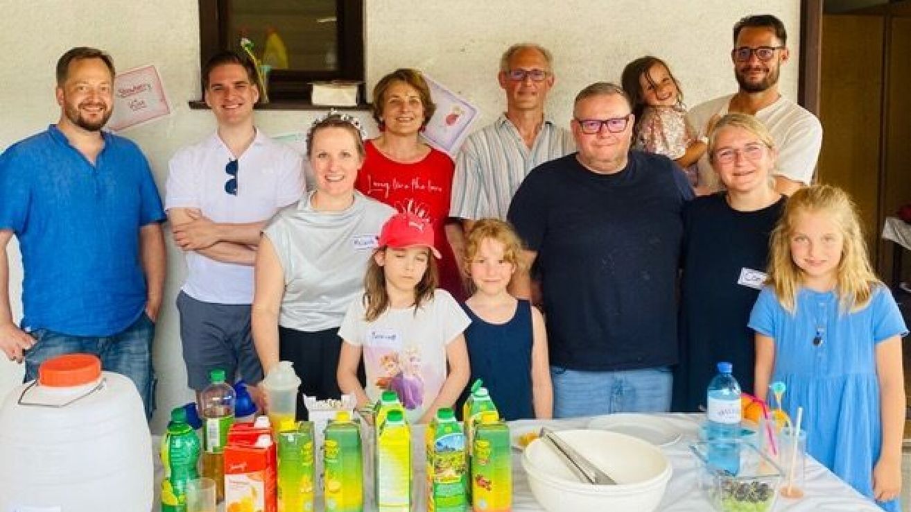 Ein Familientreffen mit mehreren Erwachsenen und Kindern, die lächeln und für ein Foto posieren. Zwei junge Mädchen sind vorne. Flaschen und Becher stehen auf dem Tisch.