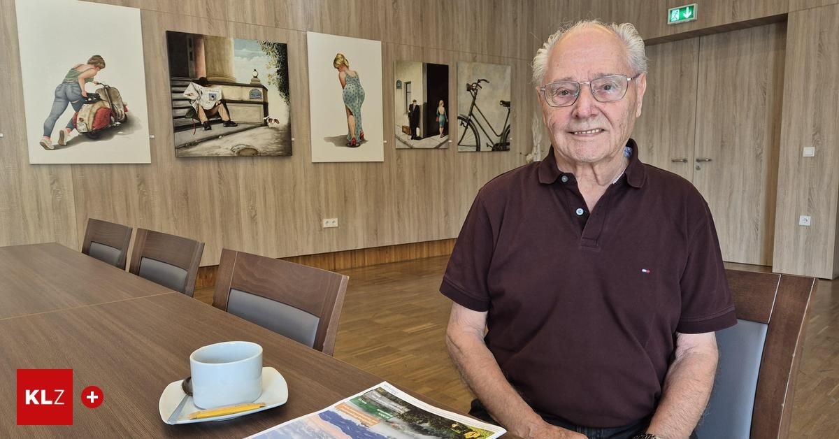 Ein älterer Mann in einem braunen Poloshirt sitzt lächelnd an einem Tisch in einer Kunstgalerie. Auf dem Tisch stehen eine Tasse, eine Untertasse und ein Bleistift. Die Holzwände haben verschiedene Gemälde.