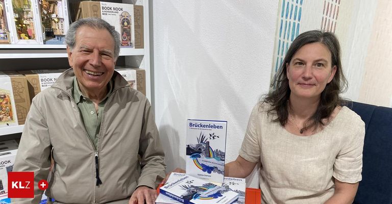 Ein Mann und eine Frau sitzen lächelnd an einem Tisch mit Büchern. Dahinter eine weiße Wand mit Bücherregalen.