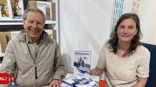 Ein Mann und eine Frau sitzen lächelnd an einem Tisch mit Büchern. Dahinter eine weiße Wand mit Bücherregalen.