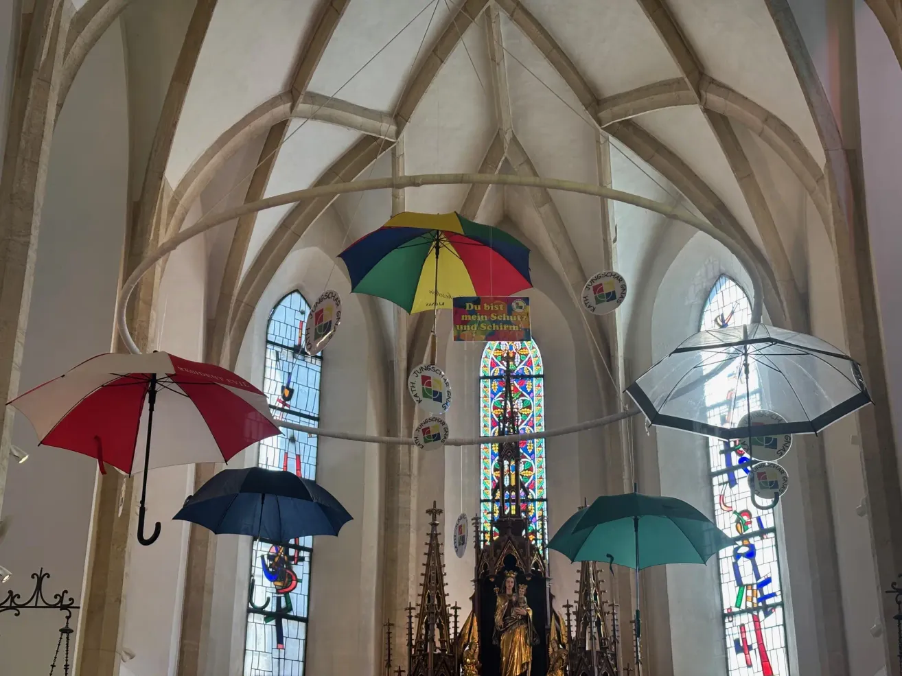 Ein Kircheninneres mit bunten Regenschirmen, die von der Decke hängen. Es gibt drei mehrfarbige Regenschirme und zwei klare. Eine Statue einer Frau, die ein Kind hält, befindet sich unter den Regenschirmen. Es gibt Buntglasfenster mit komplizierten Designs.