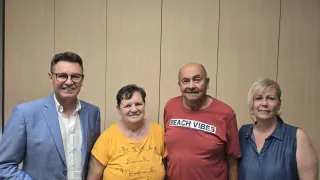 Vier Personen, zwei Männer und zwei Frauen, stehen zusammen vor einer beigen Wand. Ein Mann trägt einen blauen Anzug, der andere ein rotes T-Shirt. Die Frauen tragen gelbe und blaue Blusen. Sie alle scheinen zu lächeln.