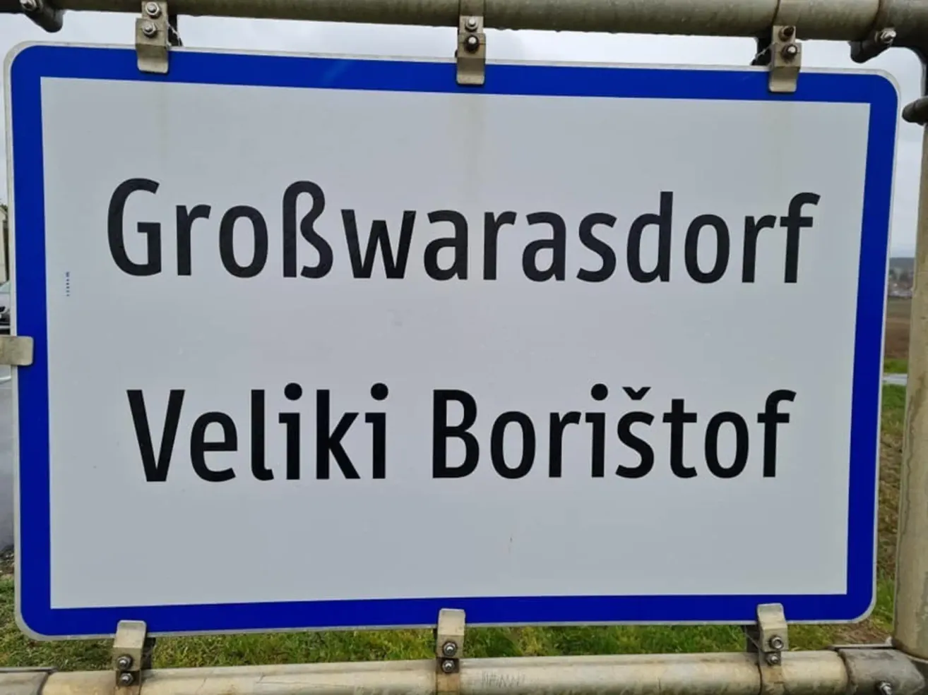 Ein weißes Schild mit blauen Rändern zeigt den Namen einer Stadt in zwei Sprachen: 'Großwarasdorf' oben und 'Veliki Boristoš' unten.