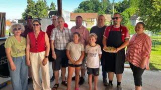Eine Gruppe von Menschen steht im Freien. Ein Mann hält eine Pizza. Die Gruppe besteht aus einer Frau in Rot, einem Mädchen mit rosa Schuhen und einem Jungen in einem weißen Hemd mit der Nummer 5. Bäume und Gebäude sind im Hintergrund.