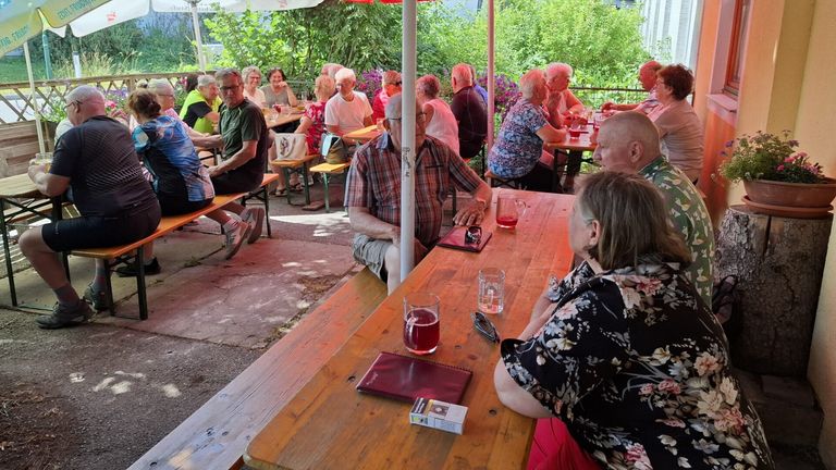 Eine Gruppe älterer Menschen sitzt um einen Holztisch in einem Garten im Freien. Einige unterhalten sich, während andere trinken. Der Garten hat grüne Pflanzen und Bäume.