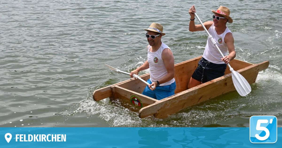 Zwei Männer paddeln ein Holzboot auf einem See, beide tragen Hüte und Sonnenbrillen. Das Boot hat ein Logo an der Seite.