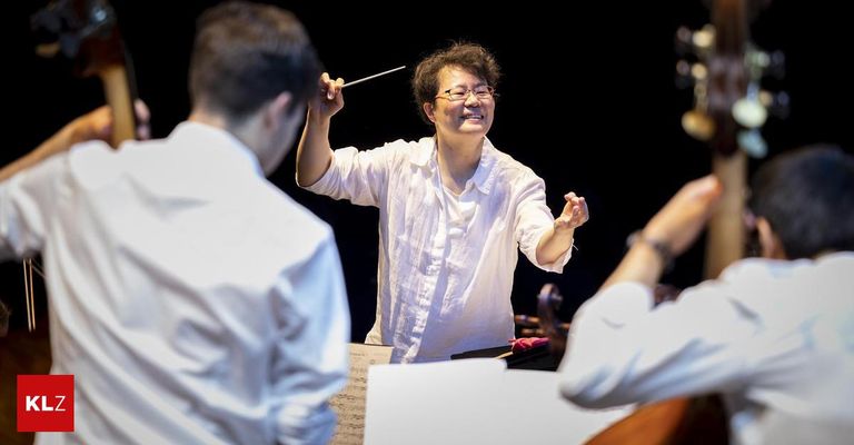 Eine Dirigentin in einem weißen Hemd führt ein Orchester an, hält einen Taktstock und lächelt, während sie zwei Musiker dirigiert, die Cello und Klavier spielen.
