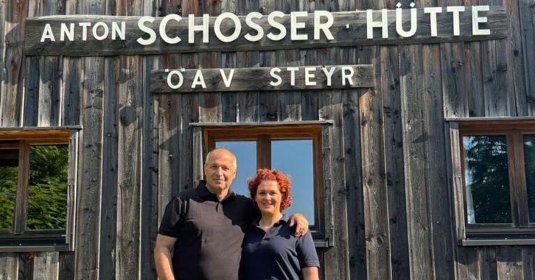 Zwei Personen stehen vor einem Holzgebäude mit einem Schild, das 'ON SCHOSSER HUT' liest. Der Mann trägt ein schwarzes Poloshirt, während die Frau rote Haare hat und ein blaues Hemd trägt. Das Gebäude hat ein Holzfenster.