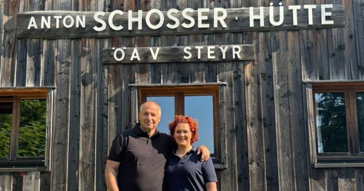 Zwei Personen stehen vor einem Holzgebäude mit einem Schild, das 'ON SCHOSSER HUT' liest. Der Mann trägt ein schwarzes Poloshirt, während die Frau rote Haare hat und ein blaues Hemd trägt. Das Gebäude hat ein Holzfenster.