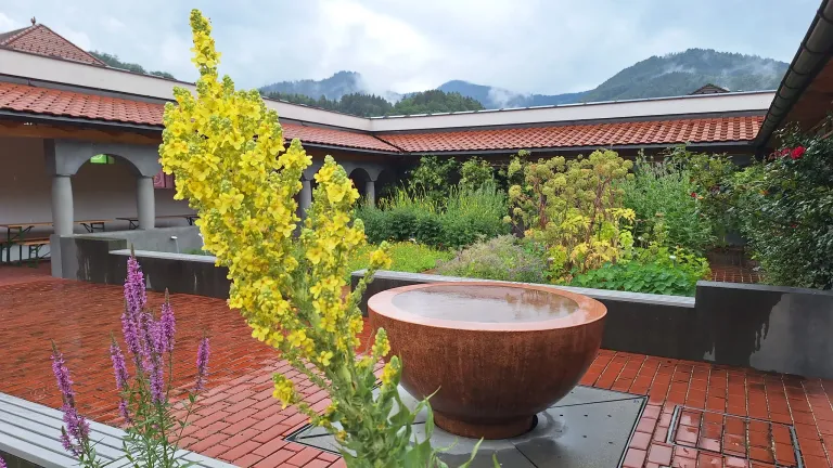 Ein Garten mit einem großen Kupferbecken, gelben Blumen und einem Gebäude mit rotem Ziegeldach im Hintergrund.