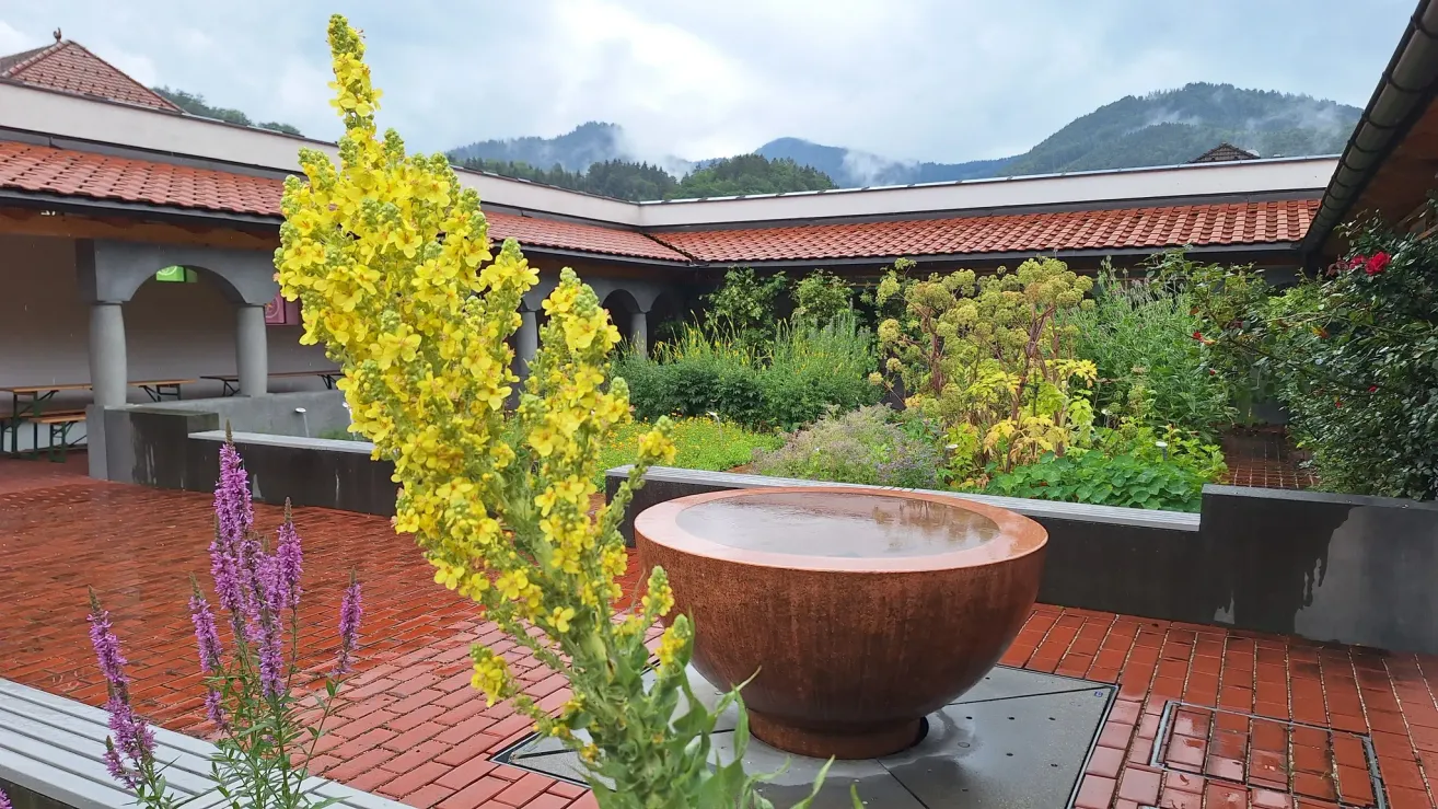 Ein Garten mit einem großen Kupferbecken, gelben Blumen und einem Gebäude mit rotem Ziegeldach im Hintergrund.