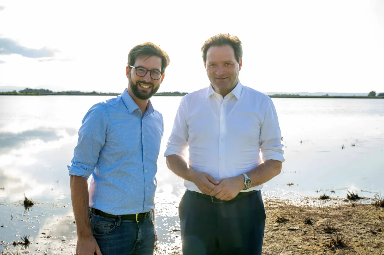 Zwei Männer stehen lächelnd am Ufer eines Gewässers. Der Mann auf der linken Seite trägt eine Brille und ein blaues Hemd, während der Mann auf der rechten Seite ein weißes Hemd und eine Uhr trägt.
